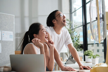 Two young Asian startup coworkers are fun blowing wind with electric fan, working in hot temperatures in a casual small business office in summer, refreshing from uncomfortable heat weather season.