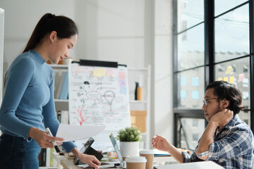 Two young Asian startup company partners discuss and brainstorm in casual small business office, marketing planning ideas, and creatively freelance work as E-commerce entrepreneur and coworker team.