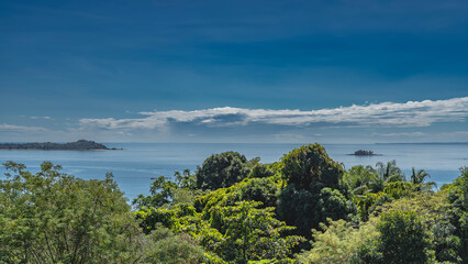 Obraz premium A tiny island is visible in the ocean. Lush green vegetation in the foreground. Blue sky, clouds. Madagascar. Nosy Komba 