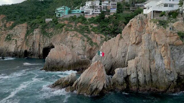 Aerial reverse shot moving away from the Mexican flag positioned near the cliffs of La Quebrada, Acapulco, revealing the expansive seascape