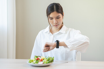 Intermittent fasting, diet healthy asian young woman dietary watching wrist watch, temptation hungry of brunch food in morning on table at home, waiting time to eat salad but not yet time first meal.