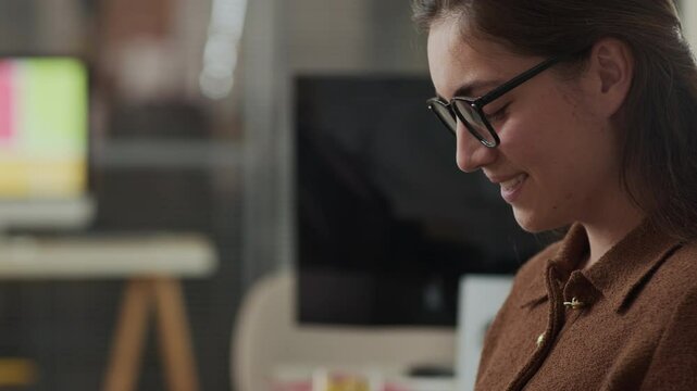 Tilt up shot of smiling young Caucasian woman wearing glasses and brown polo sweater interacting with her phone while sitting on blurred office backdrop during work day