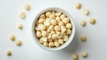 Raw macadamia nuts in a white bowl surrounded by scattered nuts on a clean, bright backdrop, showcasing their smooth, light-colored texture.