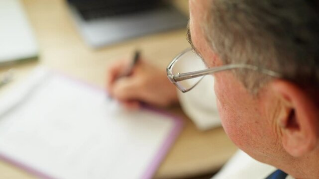Close-up of a senior general practitioner writing notes on a clipboard at a desk, wearing glasses. Concept of medical documentation, healthcare administration, or senior healthcare professional