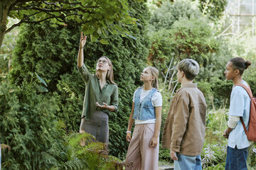 Young female teacher of biology showing trees to group of her teen students during outdoor lesson...
