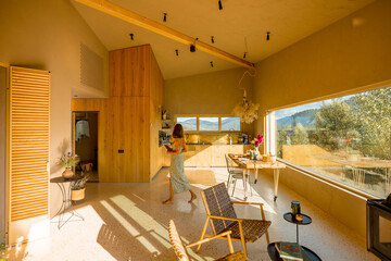 Bright and cozy kitchen with large windows showcasing mountain views. A woman in a sundress enjoys the natural light flooding into the open space