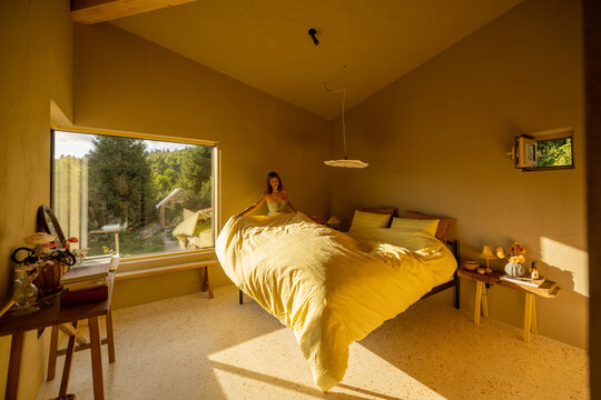 A woman making the bed with soft yellow bedding in a warmly lit bedroom. The large window offers a view of lush greenery outside, filling the room with natural light - Powered by Adobe