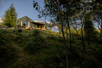 A green modern house sits on a gentle hill, surrounded by lush nature. Trees and greenery frame the scene, casting shadows in the evening light, highlighting a tranquil retreat amidst the forest.