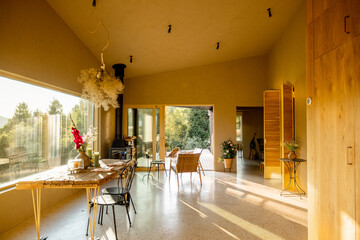 Cozy indoor space featuring a rustic wooden dining table with flowers, a wood-burning stove, and natural sunlight streaming through large windows and open doors to the deck