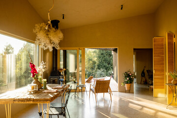 Cozy indoor space featuring a rustic wooden dining table with flowers, a wood-burning stove, and natural sunlight streaming through large windows and open doors to the deck