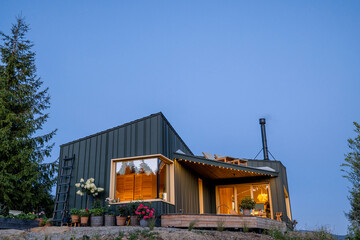 Modern cabin at dusk with soft, warm interior lighting illuminating the cozy living space. Beautiful garden in bloom complements the tranquility of the mountain surroundings