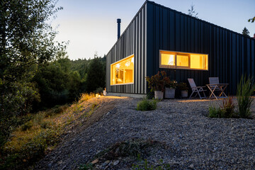 Obraz premium A modern cabin nestled in nature, illuminated warmly at dusk. Surrounded by lush greenery and trees, creating a serene escape in the wilderness
