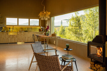Open kitchen and living area with a fireplace, rustic decor, and a large window overlooking lush greenery. Warm natural light fills the space, creating a peaceful and cozy atmosphere