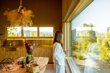 Fototapeta premium A woman stands by a large window, enjoying a peaceful morning with a cup of coffee. The rustic kitchen with warm wooden accents contrasts the lush greenery outside