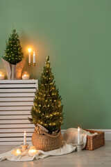 Small spruce trees with Christmas lights and other decor near green wall indoors