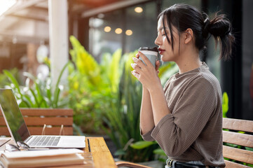 A beautiful, calm Asian woman enjoys a hot drink while working remotely from a coffee shop.