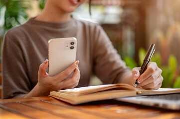 A cropped image of a woman holding her smartphone and writing in a book.