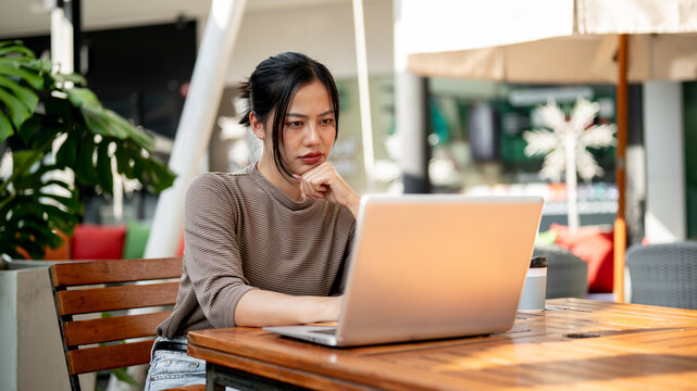 A serious, focused Asian woman reads an article on her laptop or plans her work, working remotely.