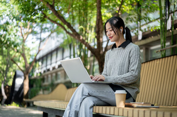 A charming Asian woman works remotely outdoors, sitting on a park bench with her laptop.