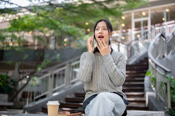 Fototapeta premium A cheerful woman sits outdoors, listens to a surprising story during a fun conversation on the phone