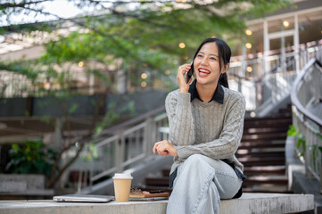 A cheerful Asian woman sits outdoors, laughing and enjoying a fun conversation on the phone.