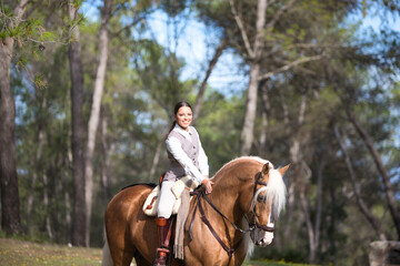 Fototapeta premium portrait of equestrian woman, young and beautiful, riding and holding the reins of her purebred brown horse with blond mane and tail. The woman is happy. Concept of love for the horse.
