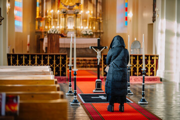 Obraz premium A woman in a black coat stands in a church during a funeral, facing the altar with lit candles and a crucifix, reflecting somber emotions..