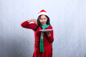 portrait of woman wearing red sweater red sweater with santa hat and scarf celebrating merry christmas, hungry look at plate carry white plate and scoop food with fork, isolated on white background