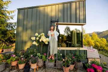 A woman stands on a ladder beside her modern mountain home, surrounded by potted plants and...