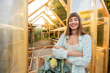 Smiling woman with a basket of fresh vegetables stands outside a greenhouse, radiating happiness and pride in her homegrown produce