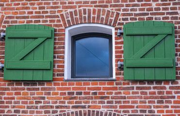 A quaint brick wall features a centered window, surrounded by green wooden shutters