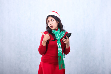 portrait of woman wearing red sweater with Santa hat and scarf celebrating merry christmas and new year, surprised to the right holding mobile phone while pointing right, isolated on white background