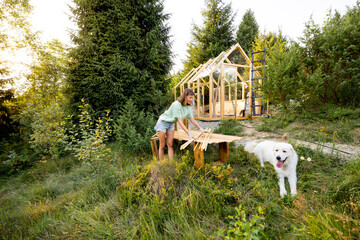 Female carpenter standing by a workbench and her large white dog in front of a partially built wooden greenhouse in her backyard. Engaged in a DIY project, surrounded by nature