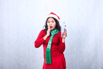 Asian young female expression wearing christmas sweater, Santa hat and scarf, very surprised hand holding white water bottle and chin, isolated on white background