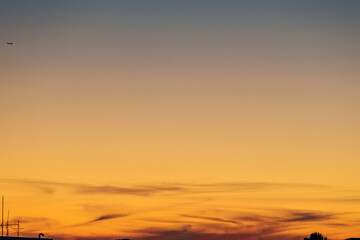 Wispy clouds swirl at the bottom of a sunset sky in transition from vivid orange to the dark hues of night. Silhouettes of an airplane, buildings, and trees add texture to the base of the scene.