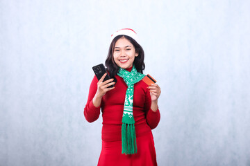 beautiful young asian woman wearing red christmas sweater with santa hat and scarf, smiling looking at camera holding debit credit and cellphone mobile phone, isolated on white background