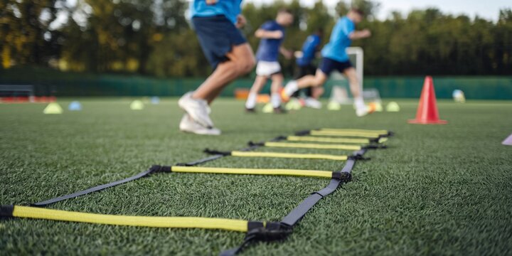 Agility Ladder Drills. A low angle, close up shot of an agility ladder lying on artificial turf with athletes in soft focus in the background. Focus is sharp on the ladder, providing a dynamic sense.