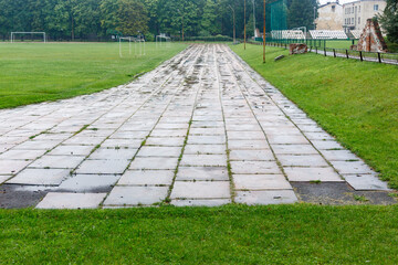 Old damaged running track in stadium with faded white lines and broken tiles, surrounded by green grass and fields. Urban sports facilities concept