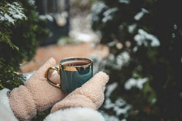 Hands in warm mittens holding a steaming cup of tea in a green mug with a golden rim, surrounded by snow-covered trees in a winter garden setting