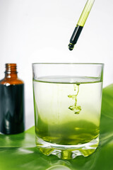 Dropper releasing green liquid into a glass of water with a leaf underneath and a brown glass bottle in the background. Represents natural ingredients, organic products