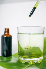 Dropper releasing green liquid into a glass of water with a leaf underneath and a brown glass bottle in the background. Represents natural ingredients, organic products.
