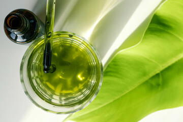 Hand holding a green dropper dripping liquid into a clear glass of water, with a green leaf and brown bottle in the background. Represents natural wellness, eco-friendly products