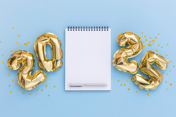 Flat lay of a blank spiral notebook with a pen, surrounded by shiny golden 2025 foil balloons on a...