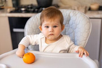 Cute little girl sitting in high baby chair with mandarin in kitchen at home. fruit healthy food kid concept. little girl daughter eating oranges at the table in the kitchen indoors.