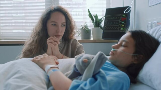 Chest up shot of young Caucasian mother holding hands together while praying for her little unconscious daughter who lying in coma in hospital bed in health centre