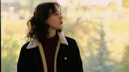 A young girl stands by a window, gazing thoughtfully at the bright outdoor scenery. This image...