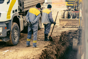 Construction workers repairing a burst water pipe under winter conditions. Excavator and truck on a muddy construction site during pipeline maintenance work.