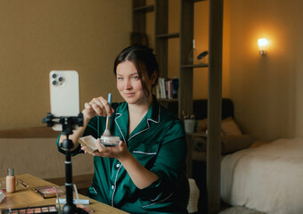 Plus size woman applying makeup on face with brush while recording vlog on smartphone for social...