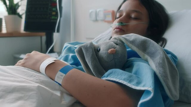 Side slowmo shot of little Caucasian girl with nasal cannula sleeping in hospital bed hugging grey bunny plush while recovering from disease in child health centre