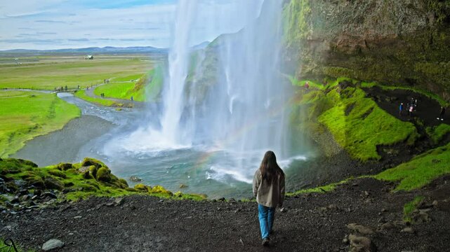 A young woman visits the tourist attraction of a volcanic glacier-fed waterfall cascading from a sheer cliff in Iceland. High Seljalandsfoss cascade, female tourist explores waterfall walk behind it. 